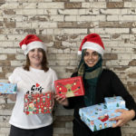 Two women in front of a white brick wall wearing red Santa hats and holding wrapped Christmas presents