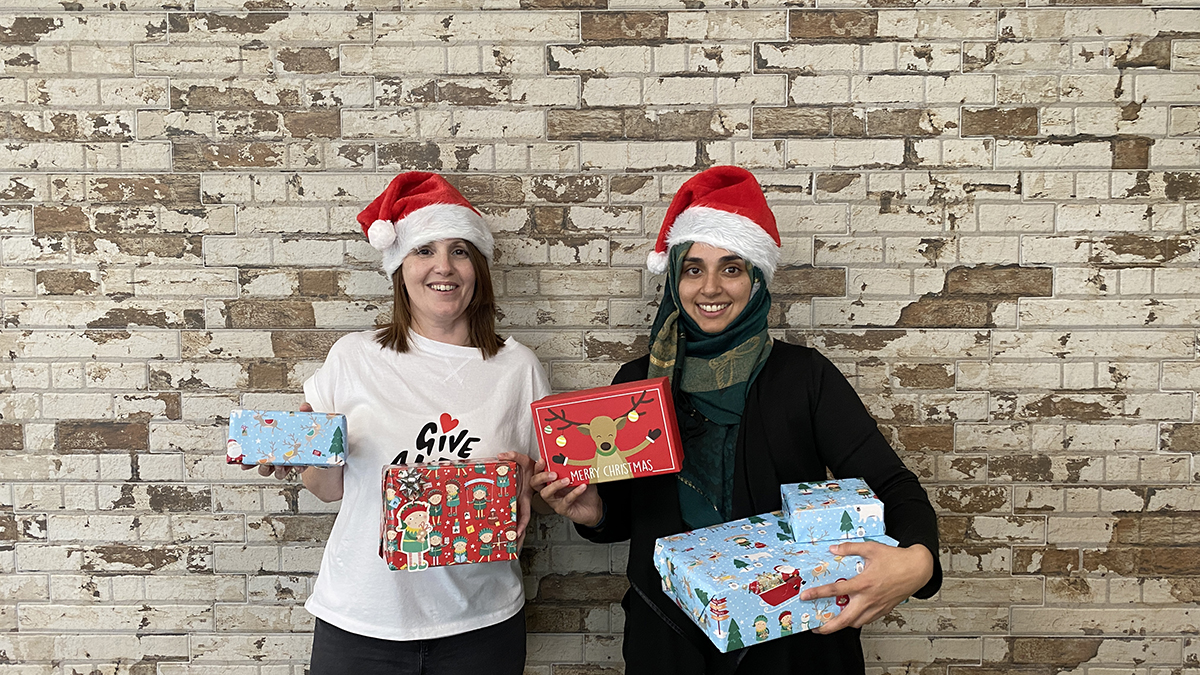 Two women in front of a white brick wall wearing red Santa hats and holding wrapped Christmas presents